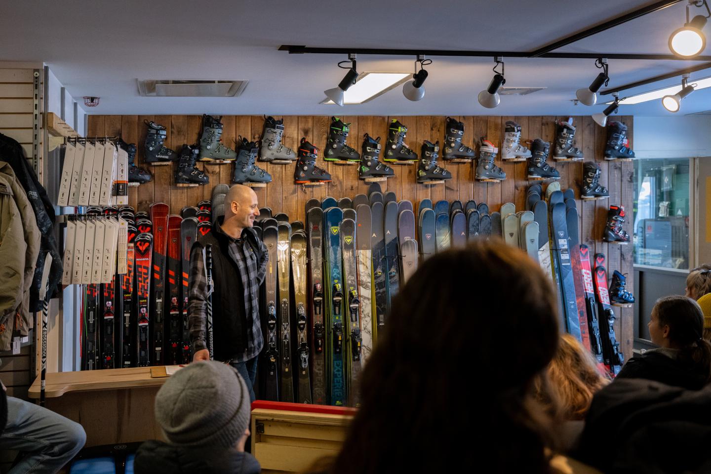 Boutique de ski avec skis et chaussures alignés au mur, homme parlant à un groupe.