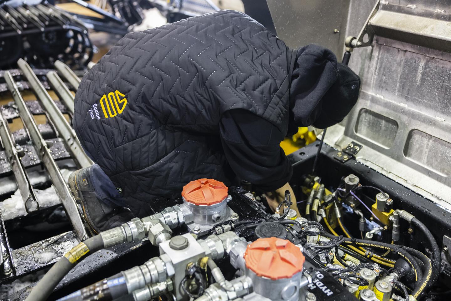 Personne réparant un moteur, vêtue d'une veste matelassée noire.