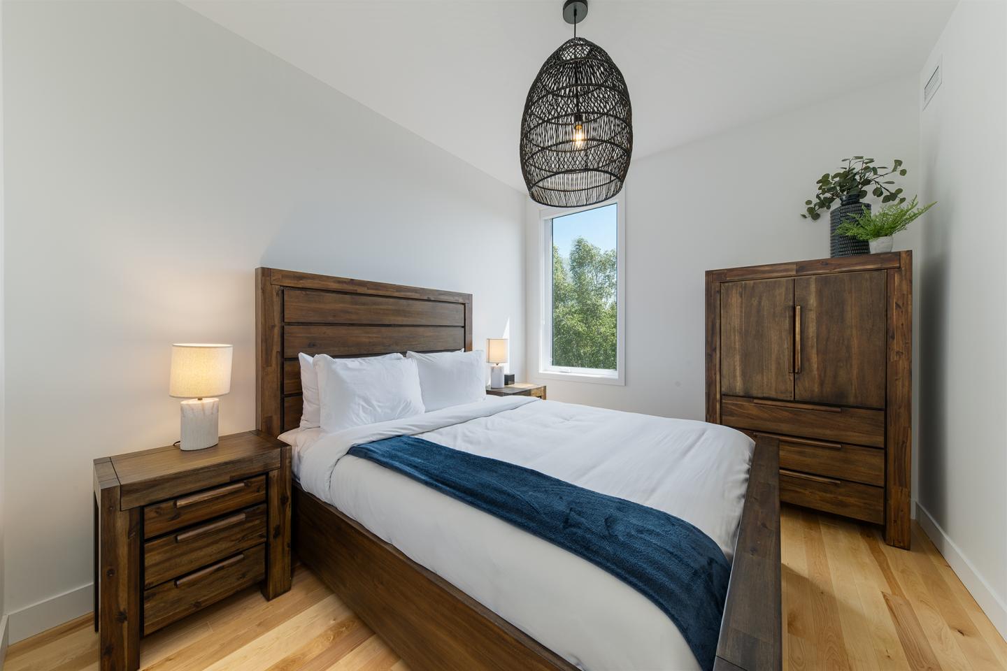 Chambre moderne avec lit en bois, armoire, et lampe allumée. Lumineuse avec une fenêtre.