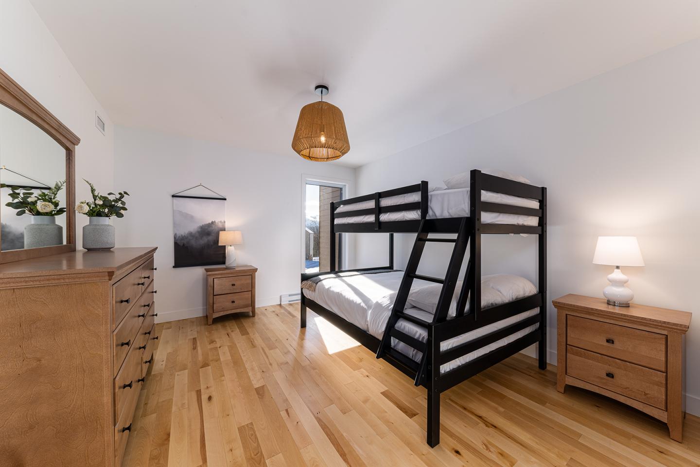Chambre avec lits superposés noirs, parquet clair, mobilier en bois, lampe et mur blanc.