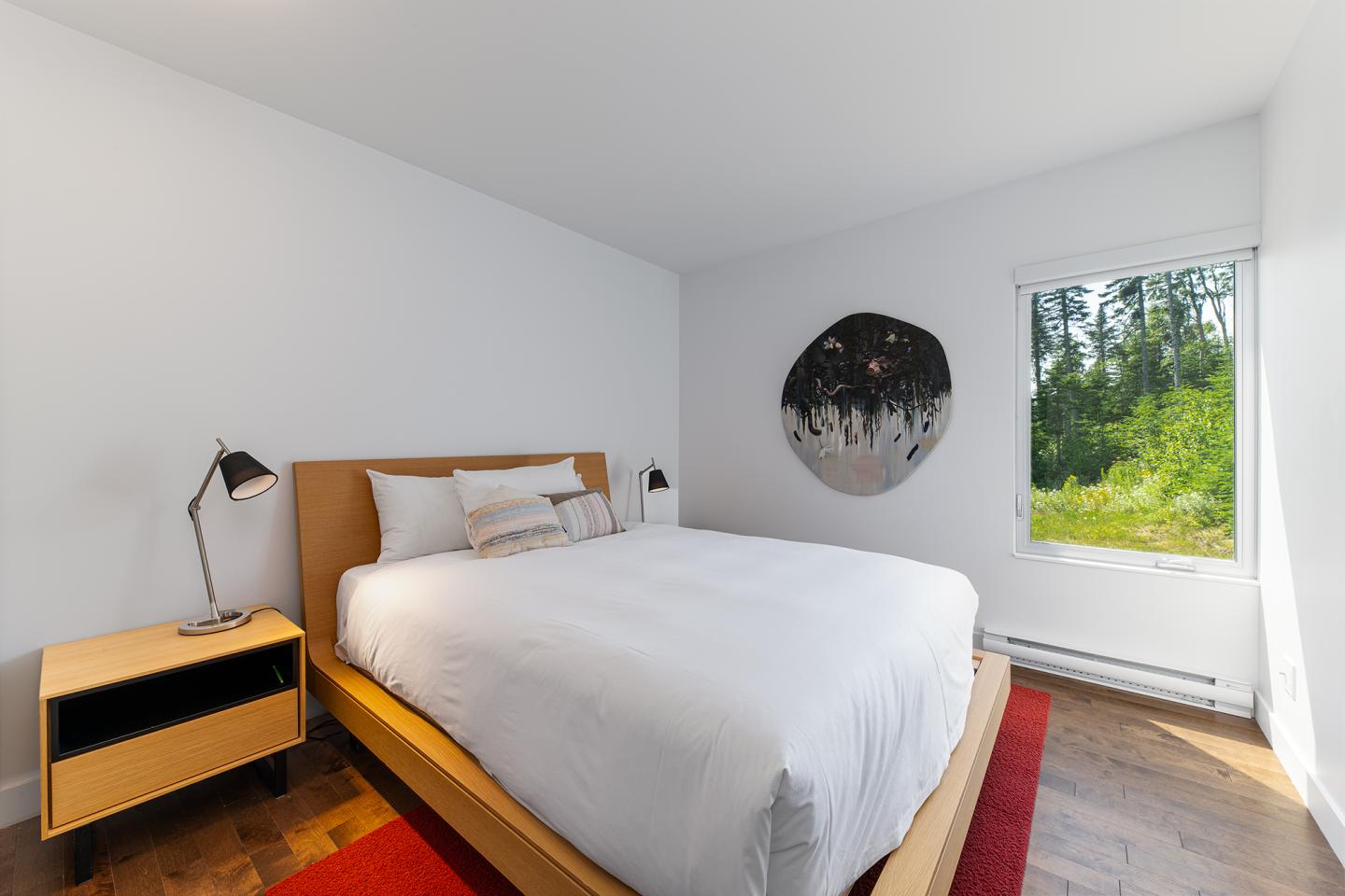 Chambre lumineuse avec lit double, mur blanc et fenêtre sur jardin.