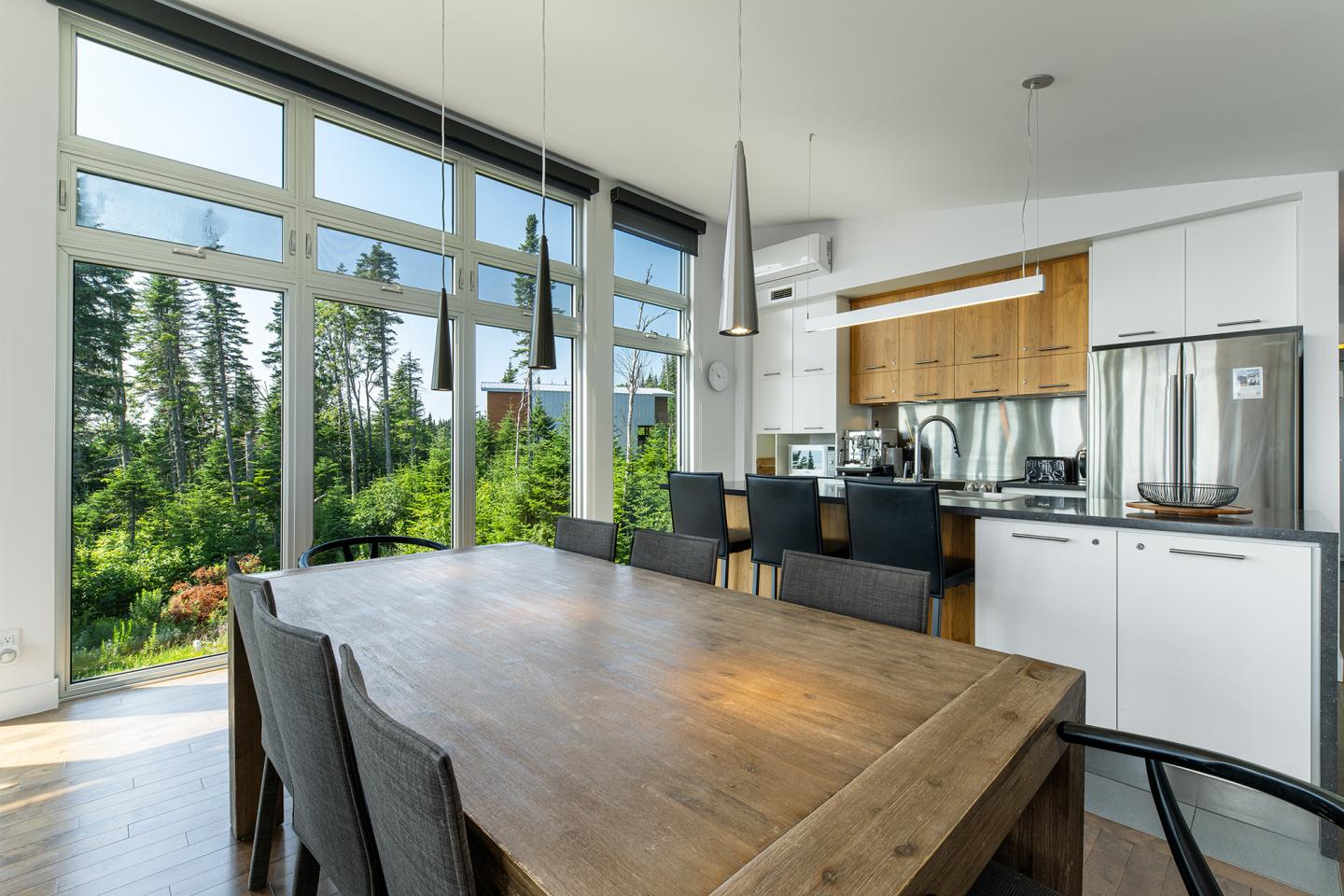 Salle à manger moderne avec table en bois, grandes fenêtres et cuisine en arrière-plan.