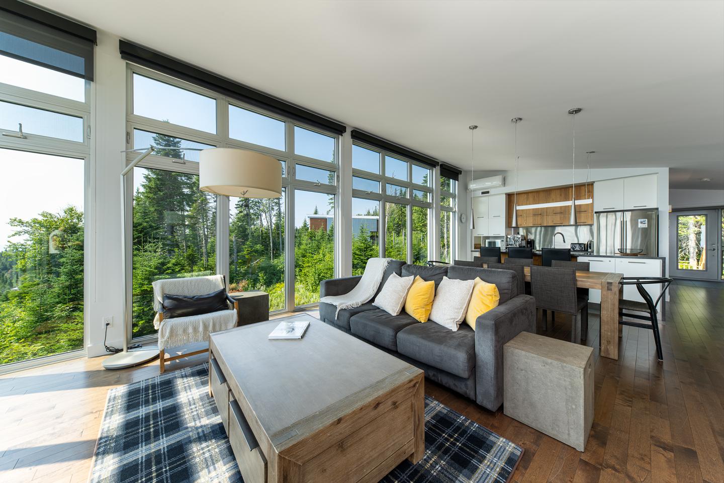 Salon lumineux avec canapé gris, coussins jaunes, grande baie vitrée, vue sur jardin verdoyant.