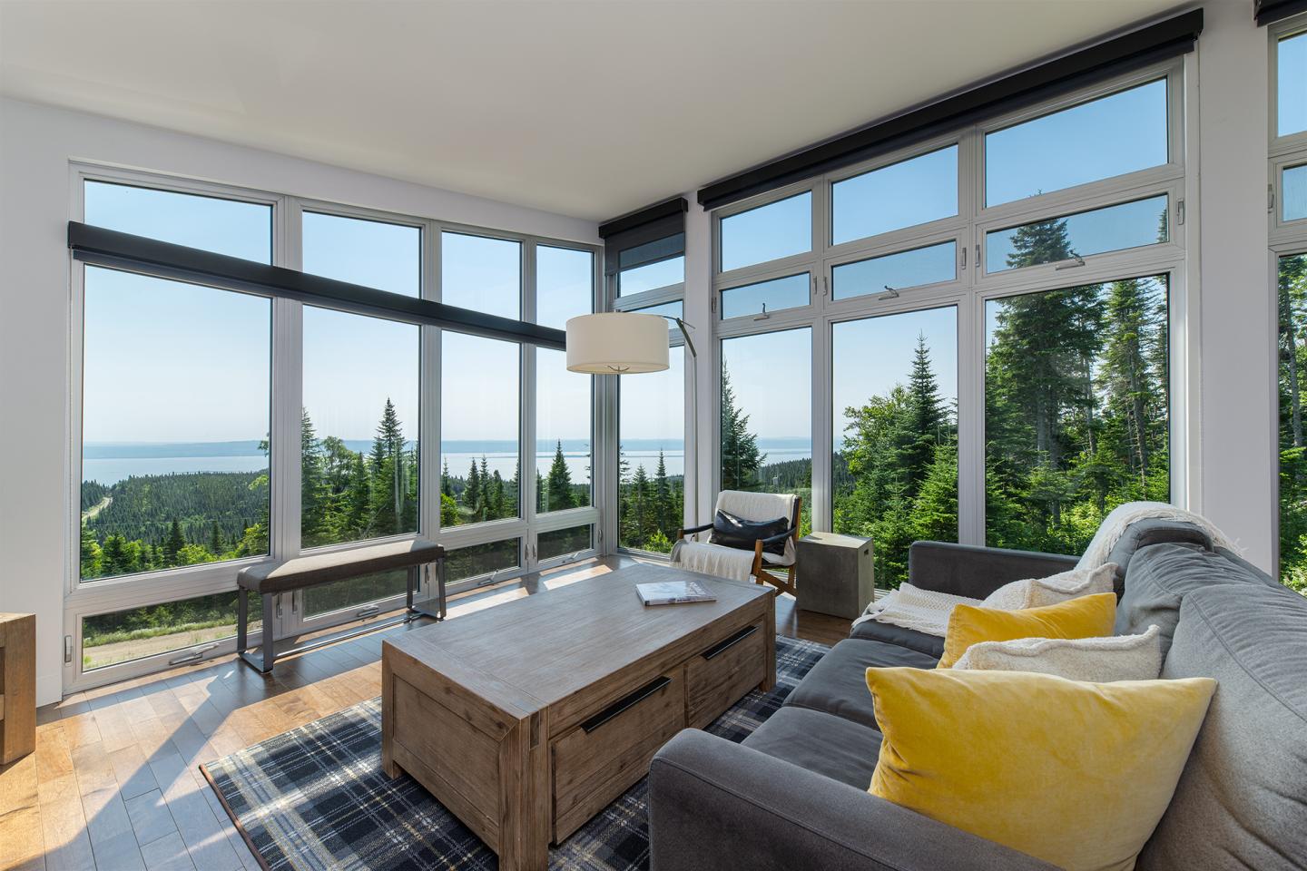 Salon moderne avec vue sur la mer et forêt, canapé gris, coussins jaunes, lumière naturelle.