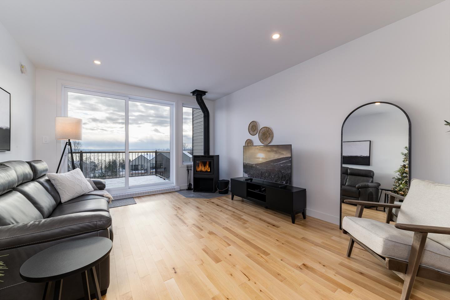 Salon moderne avec parquet, canapé noir, miroir, et vue sur mer.