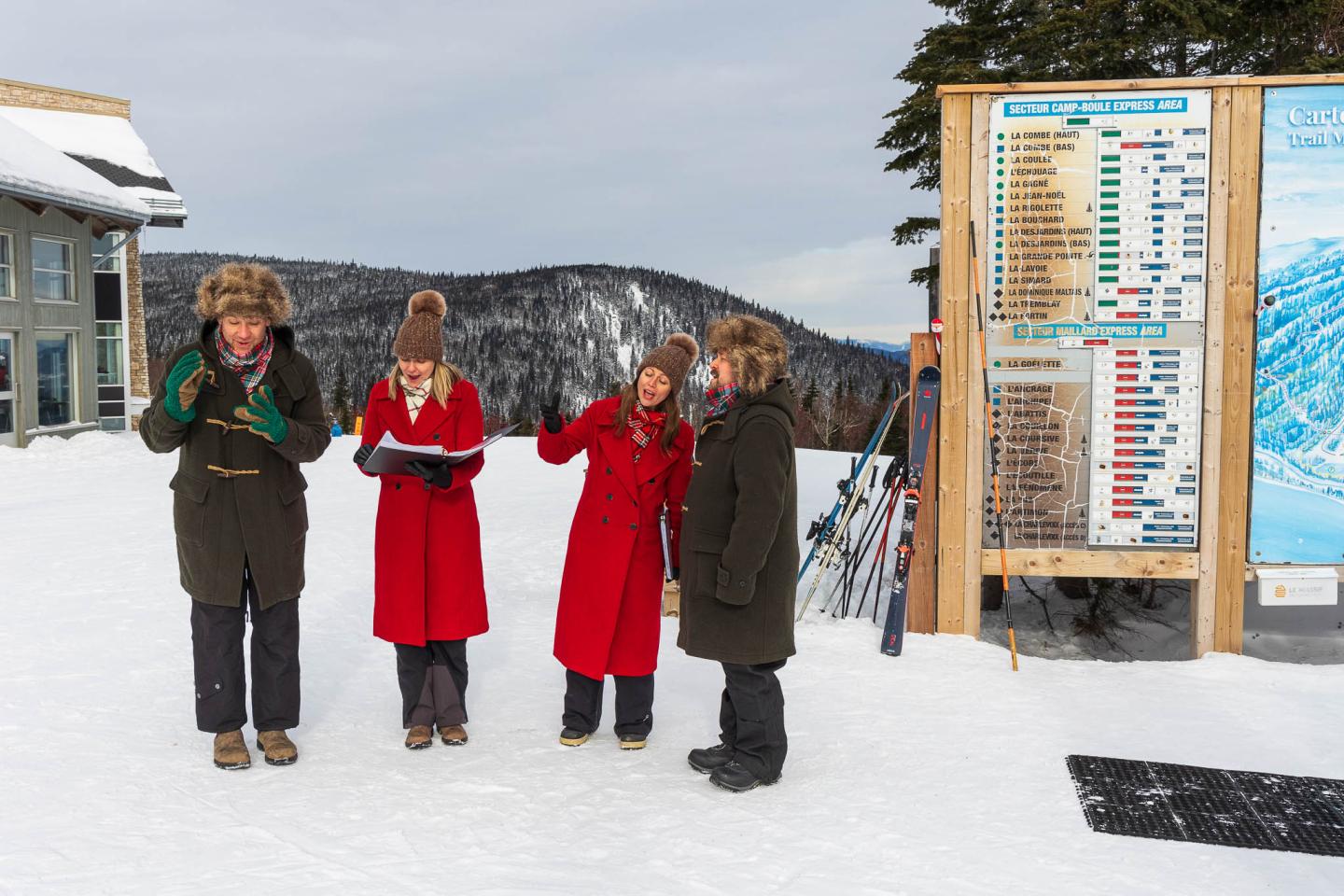 Quatre personnes en manteaux rouges et noirs devant un panneau de station de ski enneigée.
