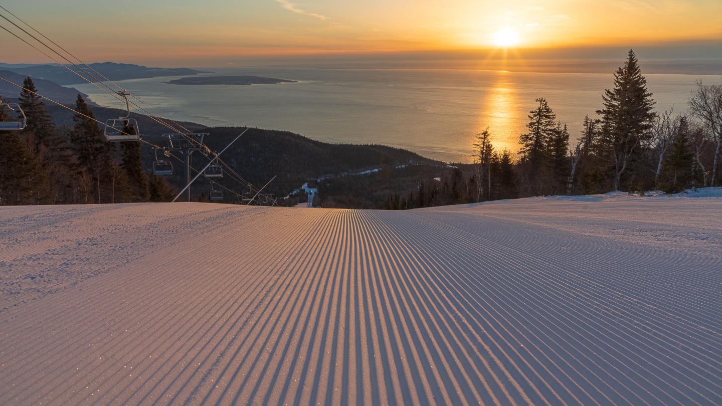 Piste de ski damée au coucher du soleil, avec lac et montagnes enneigées.