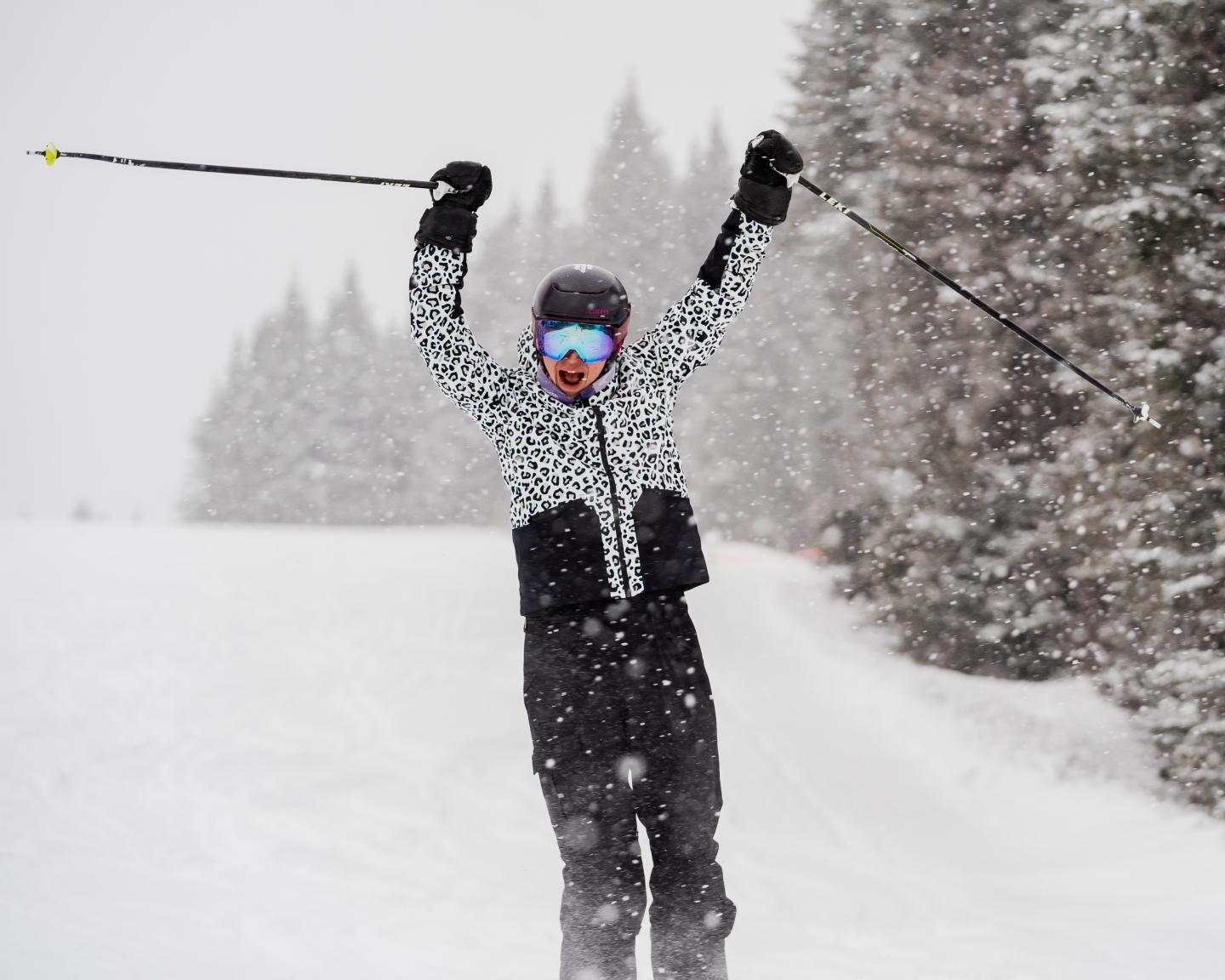 Snowboardeur en combinaison à pois, bras levés, sur une piste enneigée.