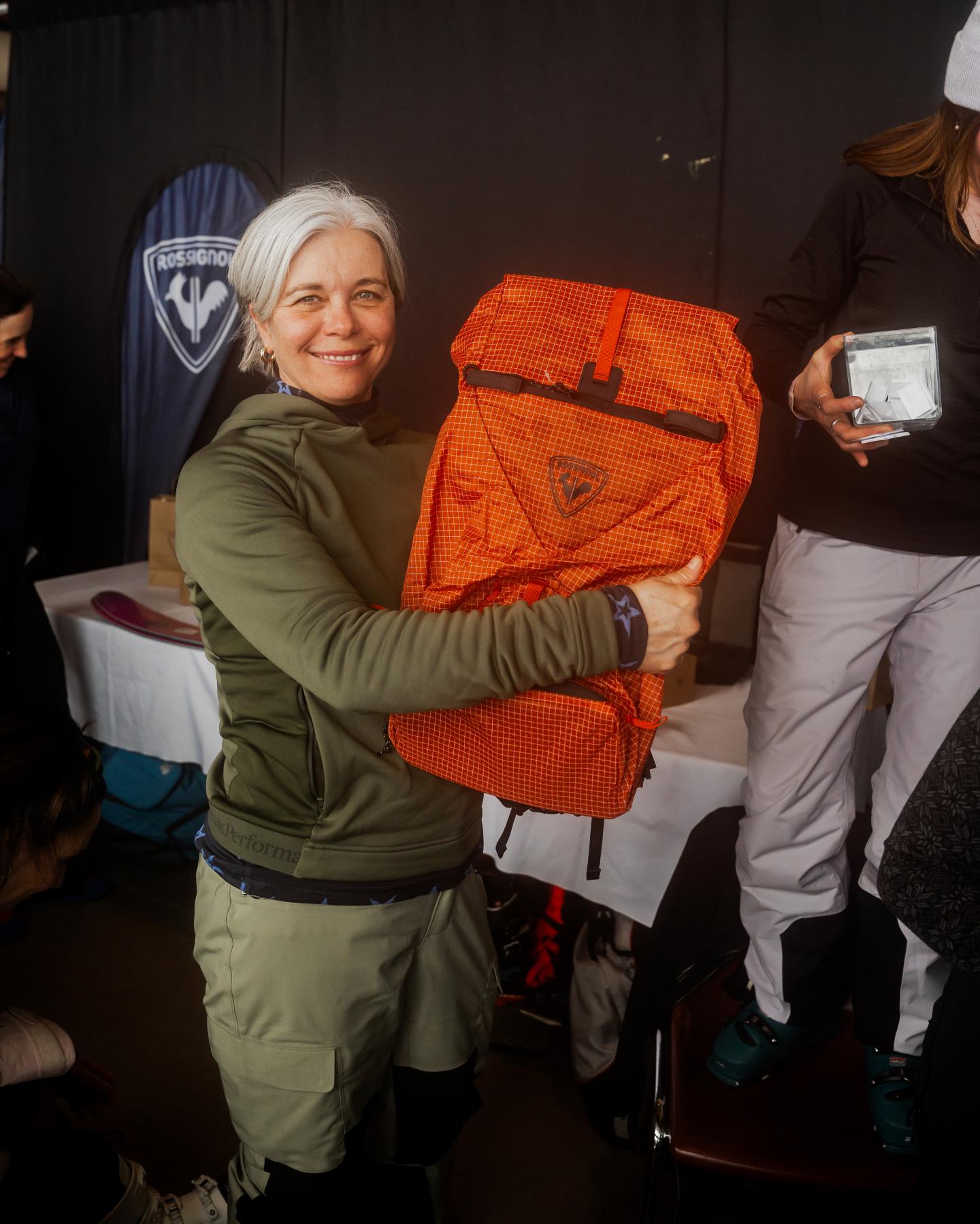 Femme souriante tenant un grand sac orange, entourée de personnes.
