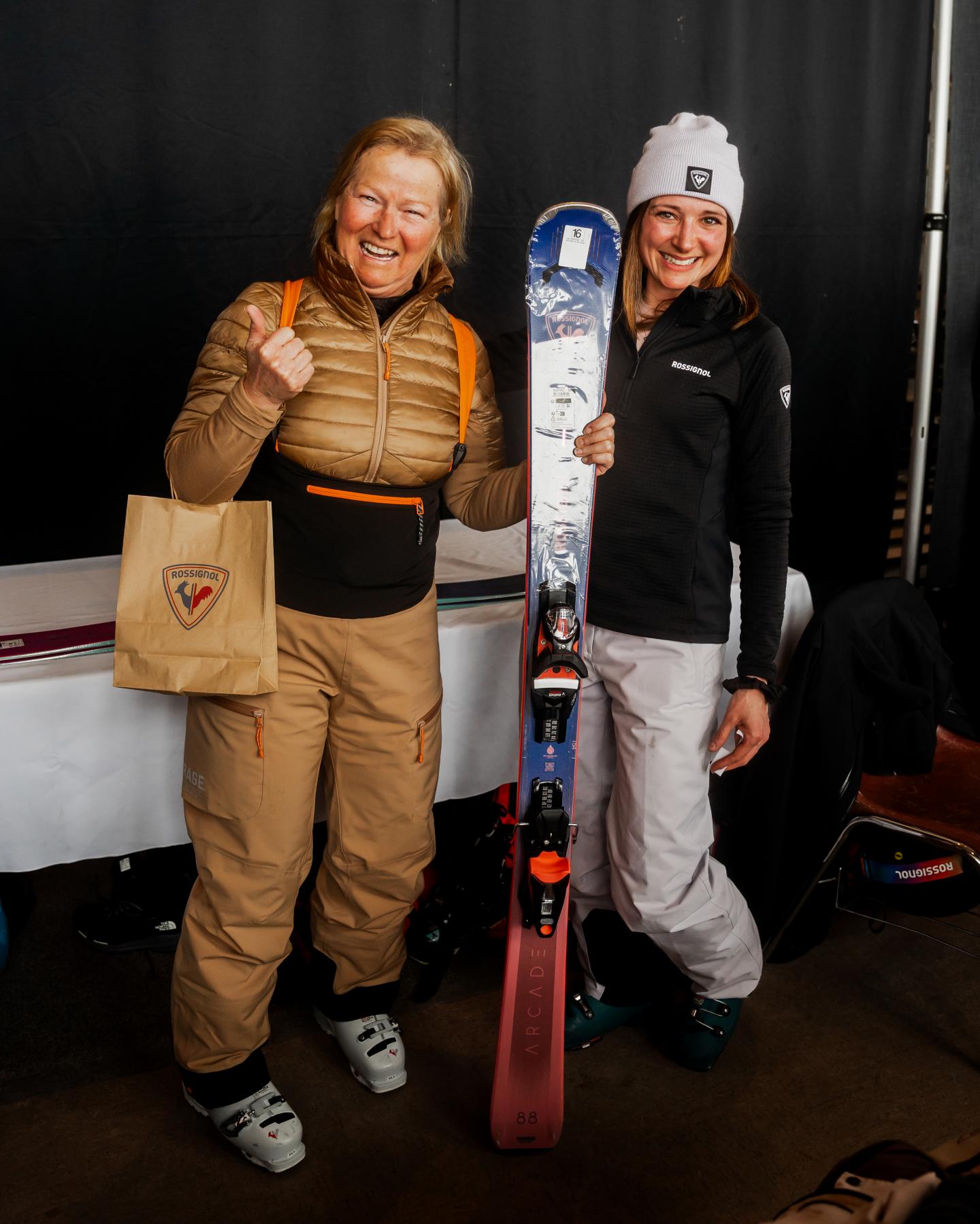 Deux skieurs souriants en tenue d’hiver, tenant des skis et des boissons.