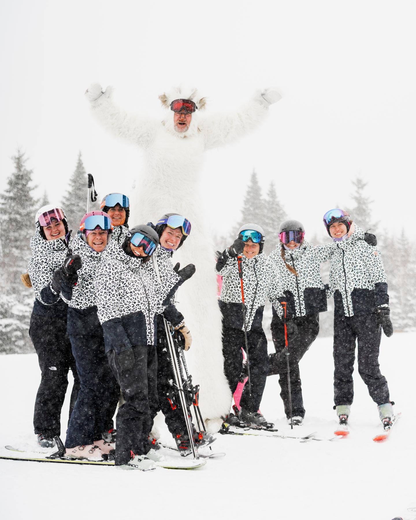 Groupe de skieurs souriants sur une piste enneigée, l’un saute bras levés.
