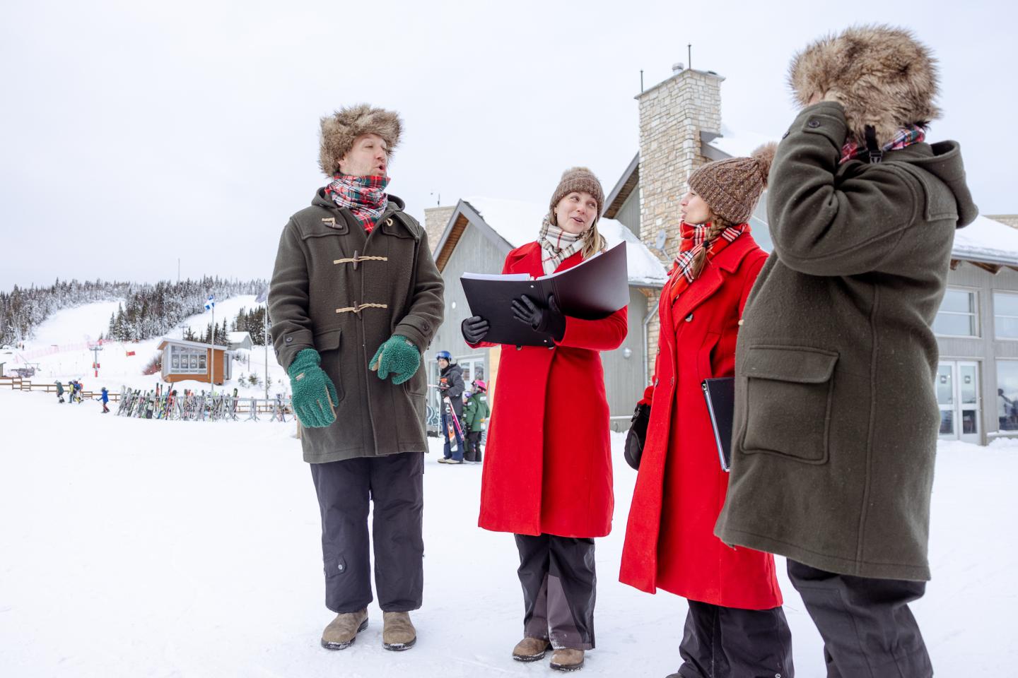 Trois chanteurs en manteaux rouges chantent dans la neige devant une femme.