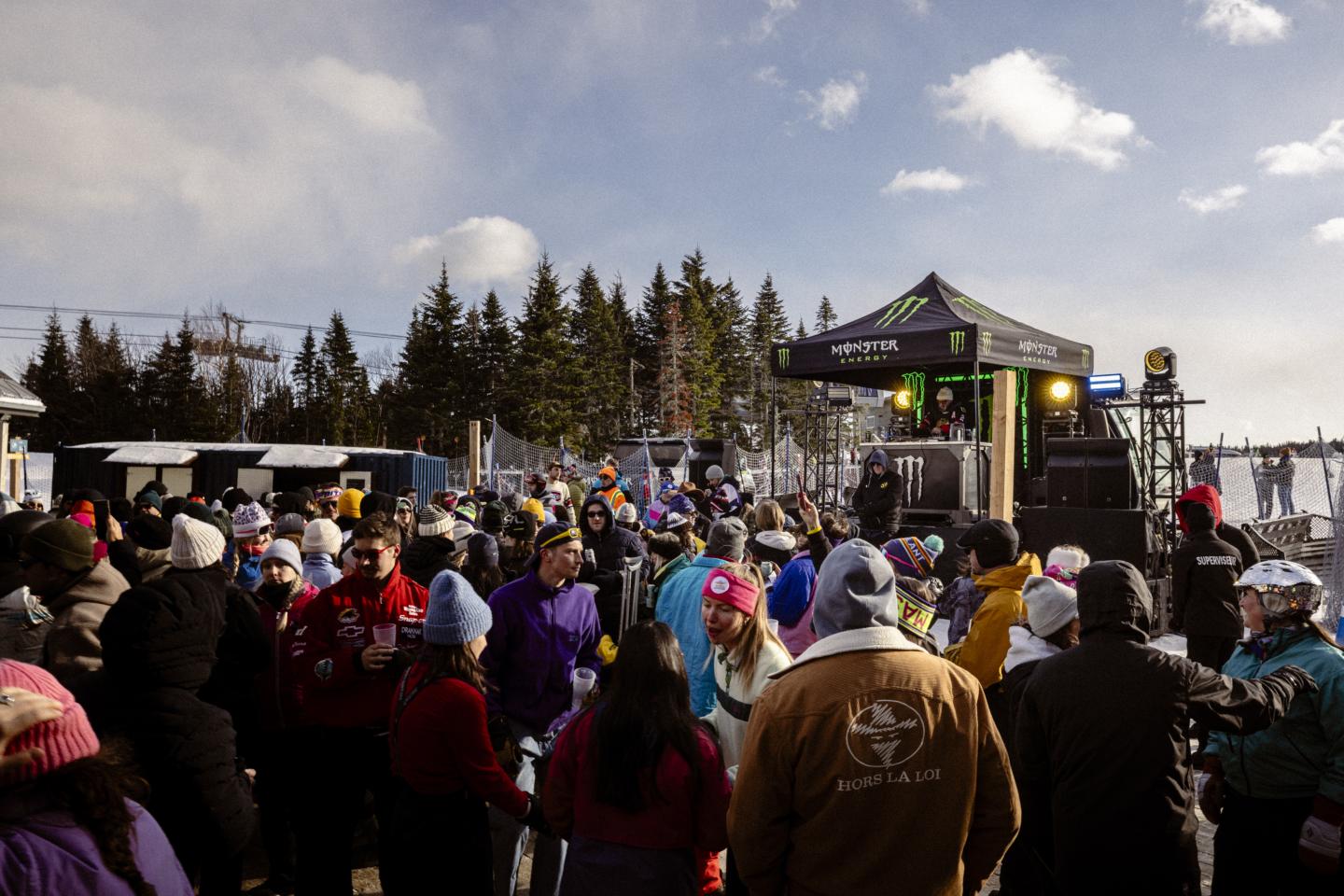 Foule en plein air devant une scène de concert, sous un ciel nuageux.