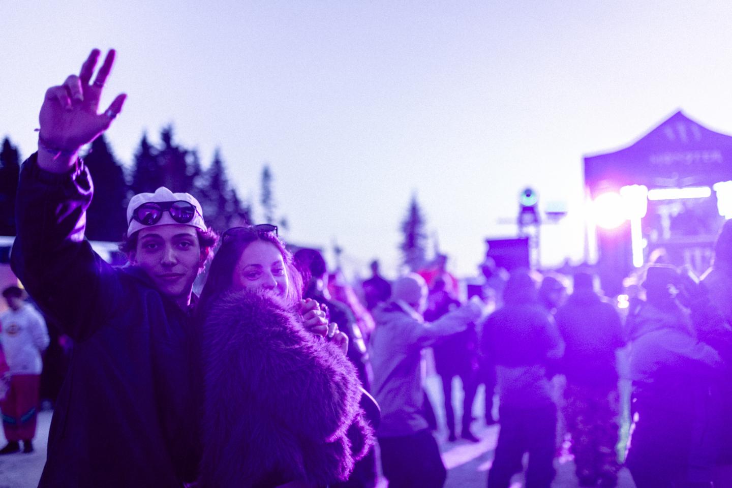 Deux amis souriants saluent la caméra lors d’un festival au crépuscule.