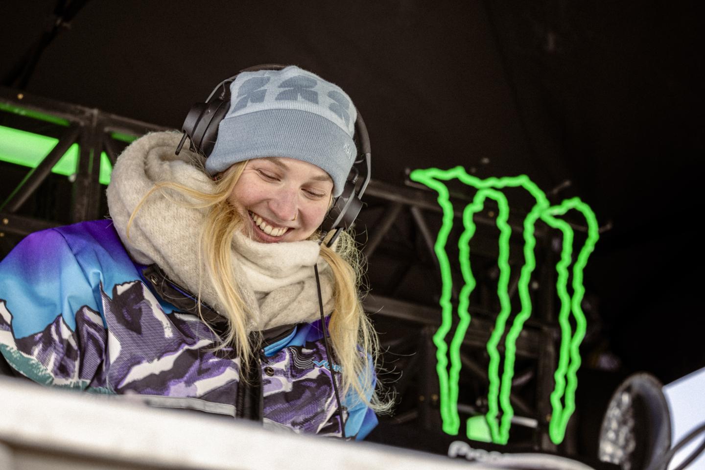 Snowboardeuse souriante en veste colorée devant le logo vert Monster Energy.
