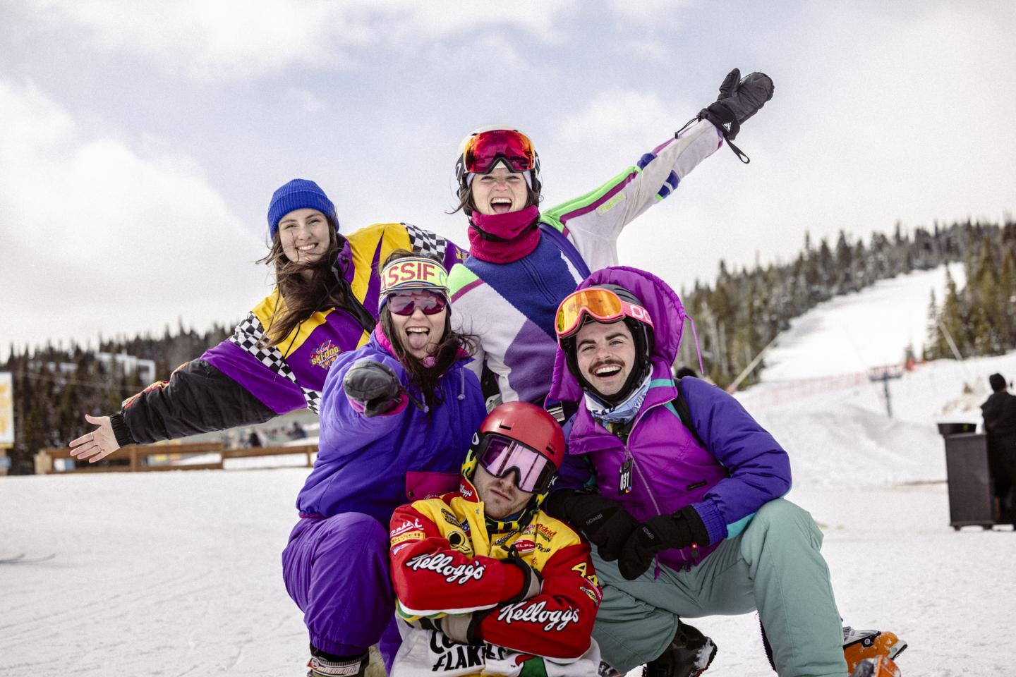Groupe souriant en vêtements de ski colorés sur une piste enneigée.