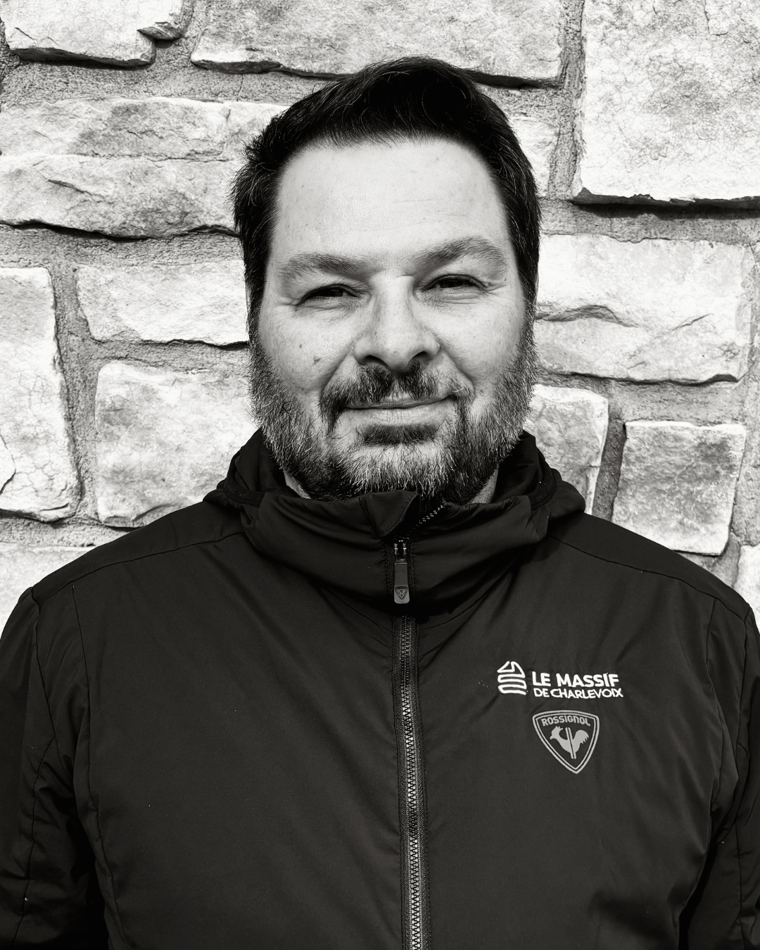 Portrait en noir et blanc d’un homme barbu devant un mur de pierre.