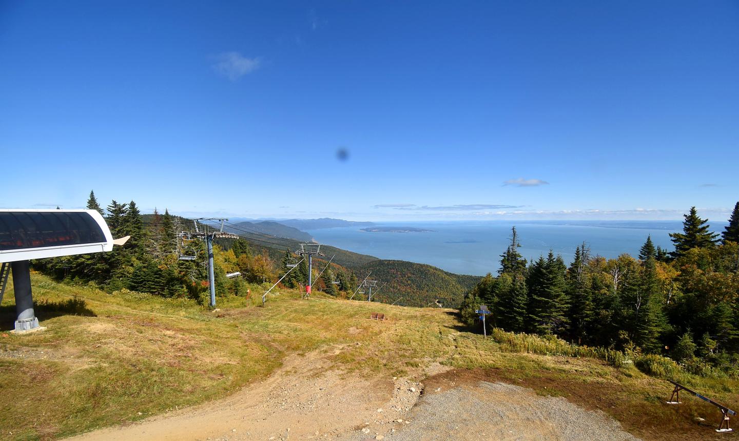 Sentier de montagne avec banc, forêt de conifères et vue sur les collines sous ciel bleu.
