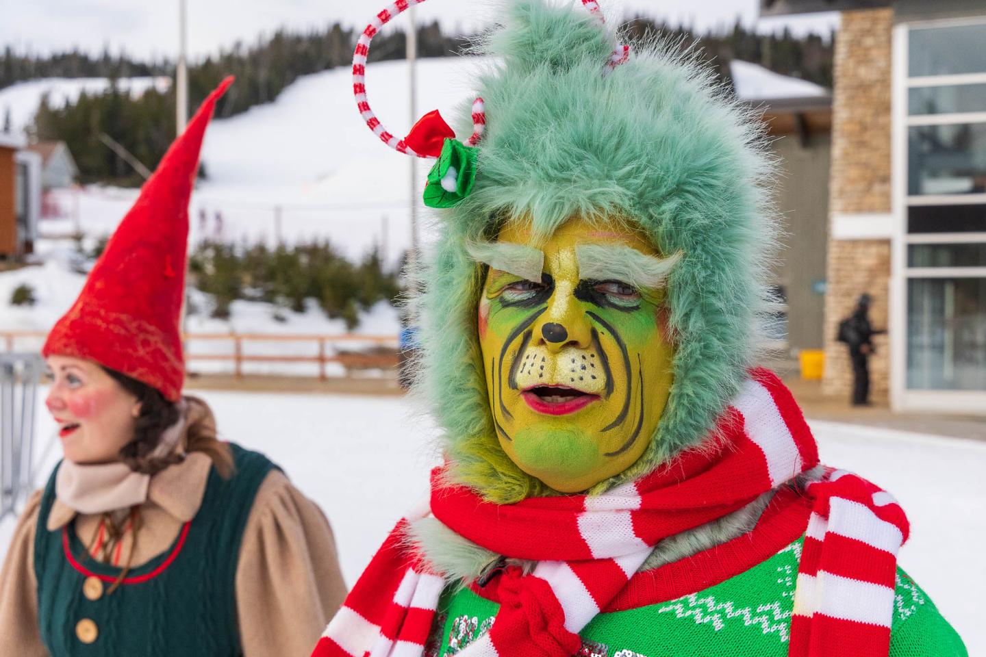 Personne déguisée en Grinch avec bonnet de Père Noël, devant une maison enneigée.