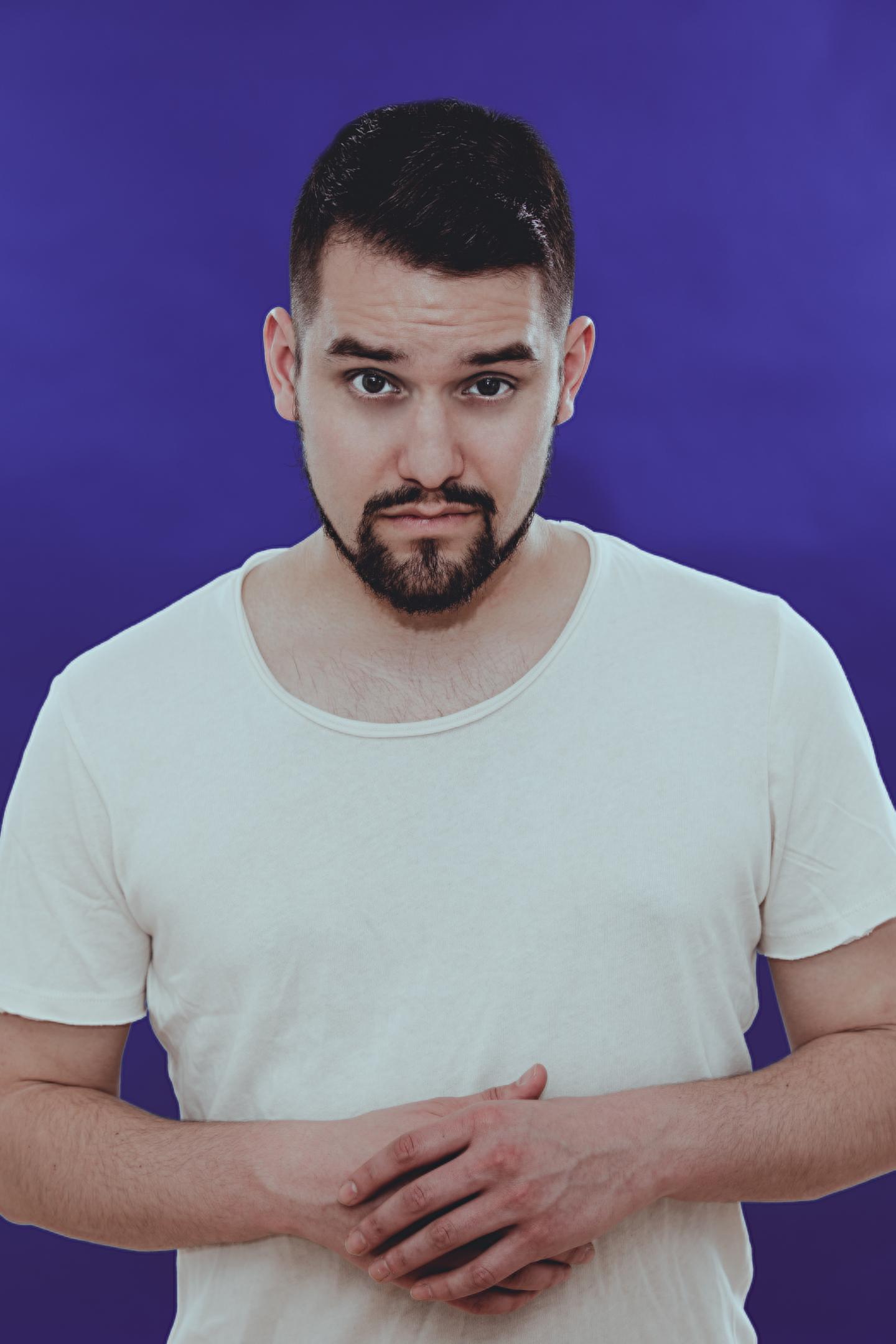 Homme barbu en t-shirt blanc, mains sur le ventre, fond violet.