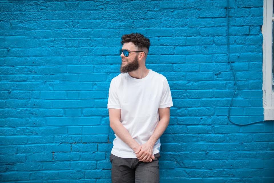 Homme barbu en t-shirt blanc devant un mur de briques bleu vif.
