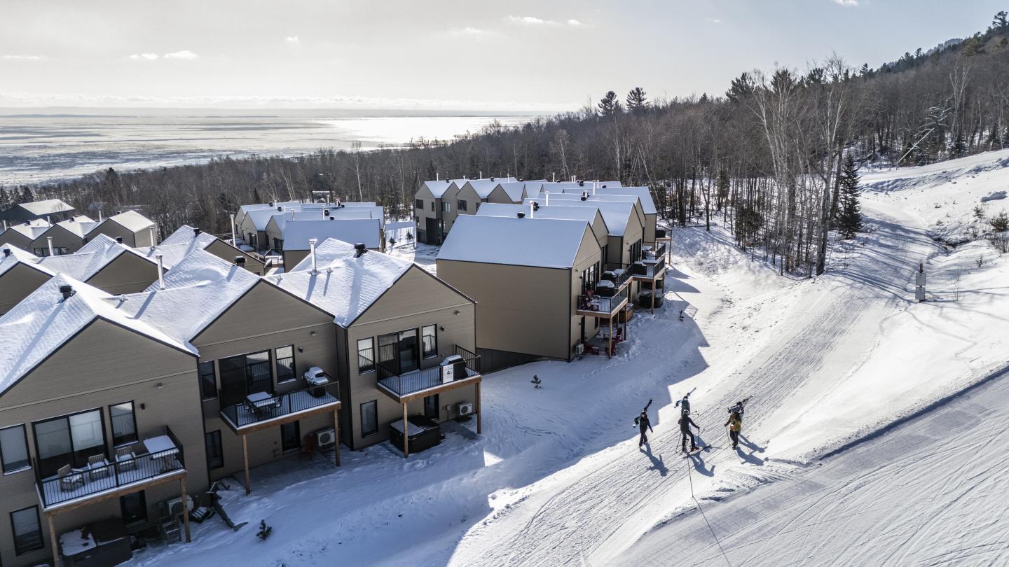 Maisons enneigées au pied d’une colline, deux skieurs sur une piste.
