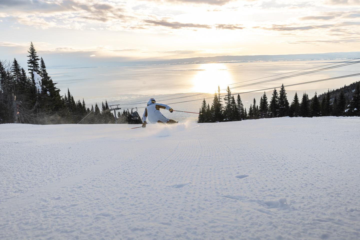 Skieurs sur une piste enneigée au coucher du soleil, bordée de sapins.